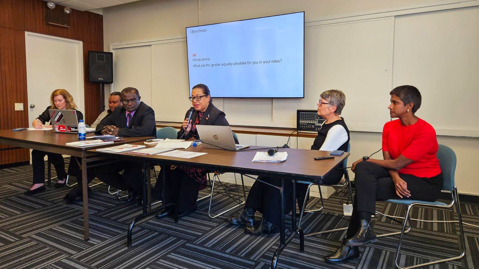 A group photo of the speakers of the event seated at the panel, from left to right - Ms. Krista Baptista Jones, Ms. Nauna Revo, Dr. Cedric Alependava, Mrs. Polotu Fakafanua-Paunga - who is holding the microphone and speaking - Ms. Joanne Crawford AM, and Dr. Gayatri Ramnath. They are seated in front of a TV that reads 'ALL: Introductions. What are the gender equality priorities for you in your roles?'