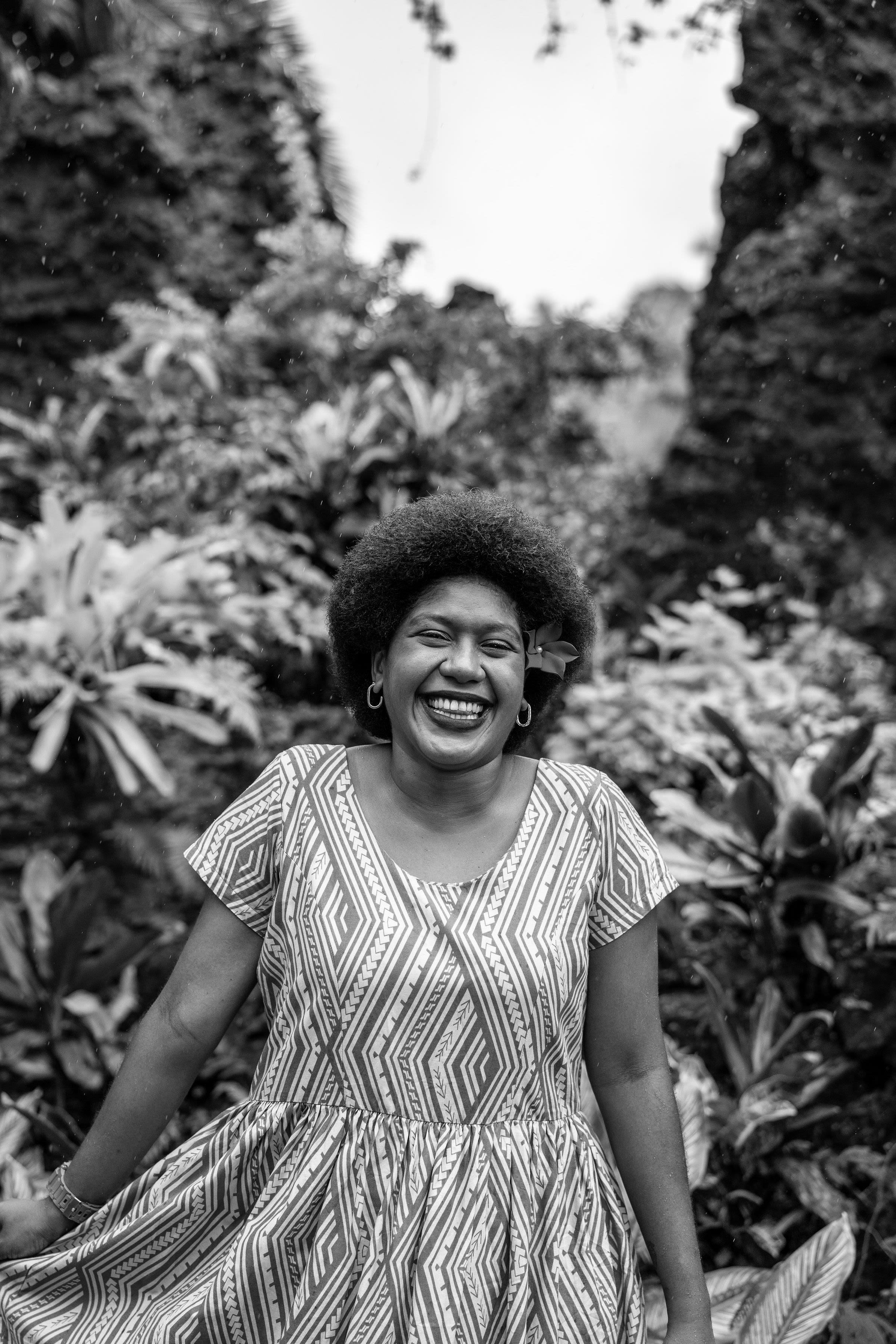 A Fijian woman smiles and has a flower in her hair. She is surrounded by plants