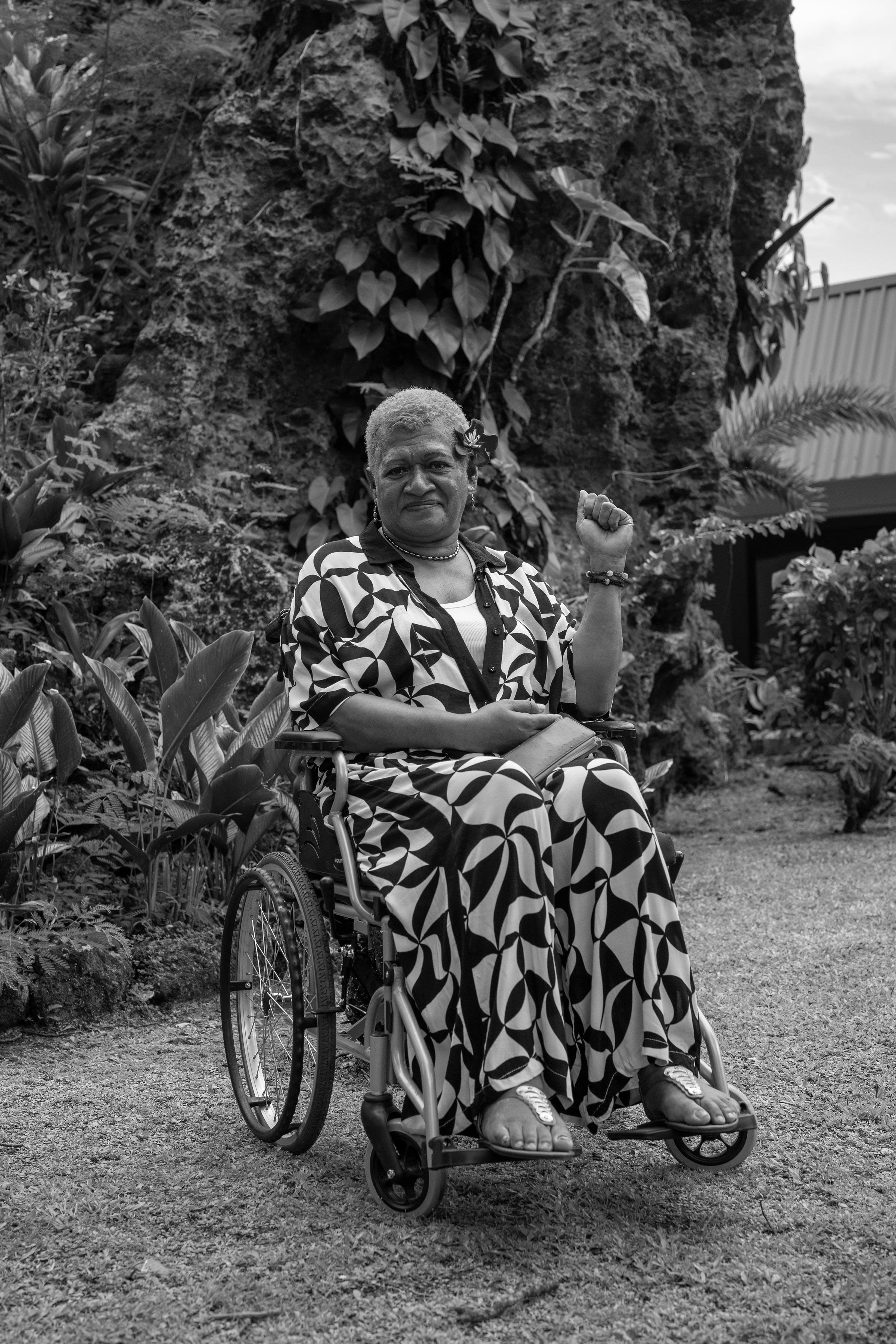A Fijian woman in a wheelchair holds up a fist and smiles, surrounded by plants