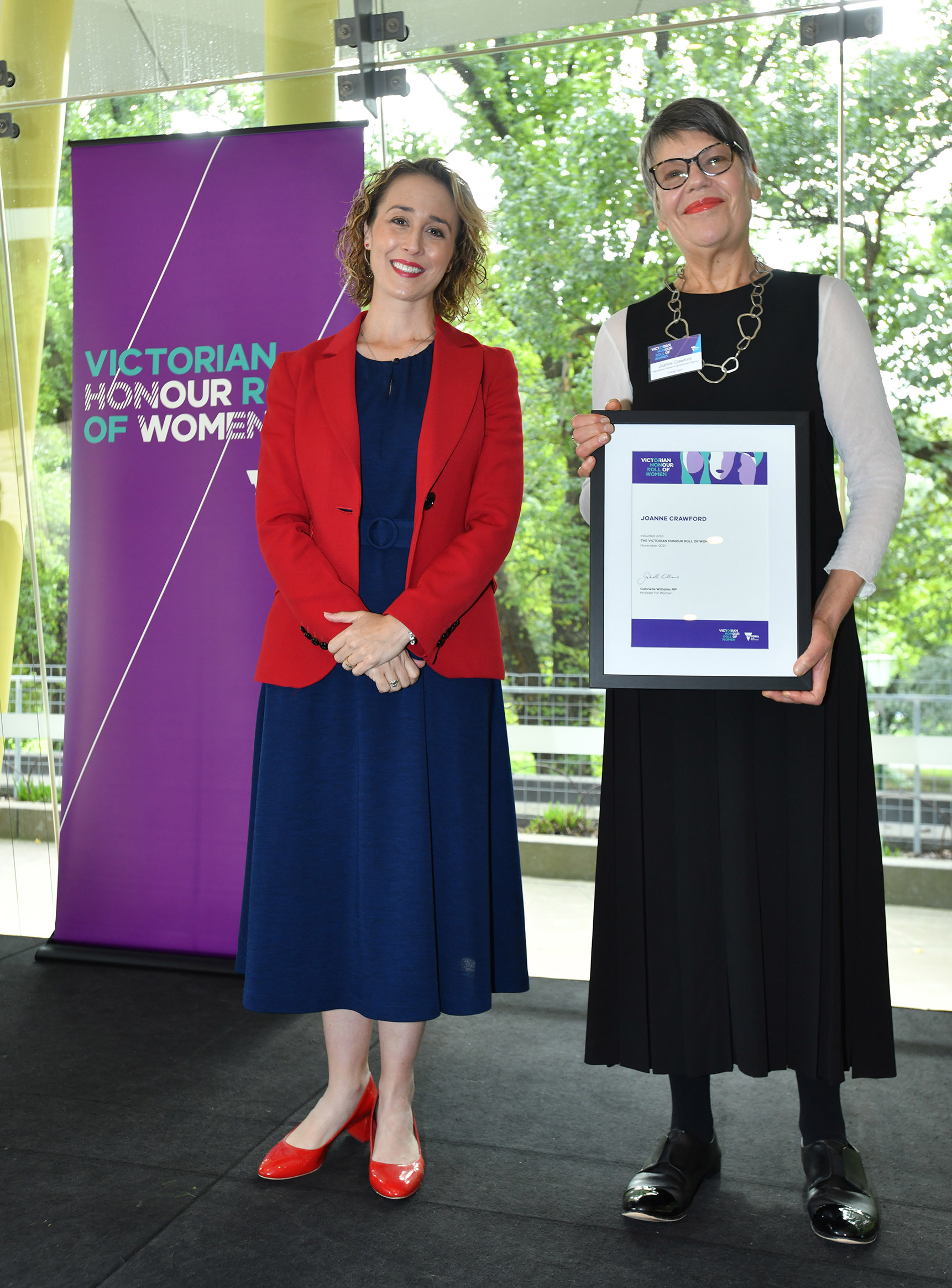 Jo a white woman wearing a long black dress is holding up an award. She is standing next to Gabrielle Williams, the Victorian Minister for Women. Gabrielle is a white woman wearing a navy dress and a red blazer.
