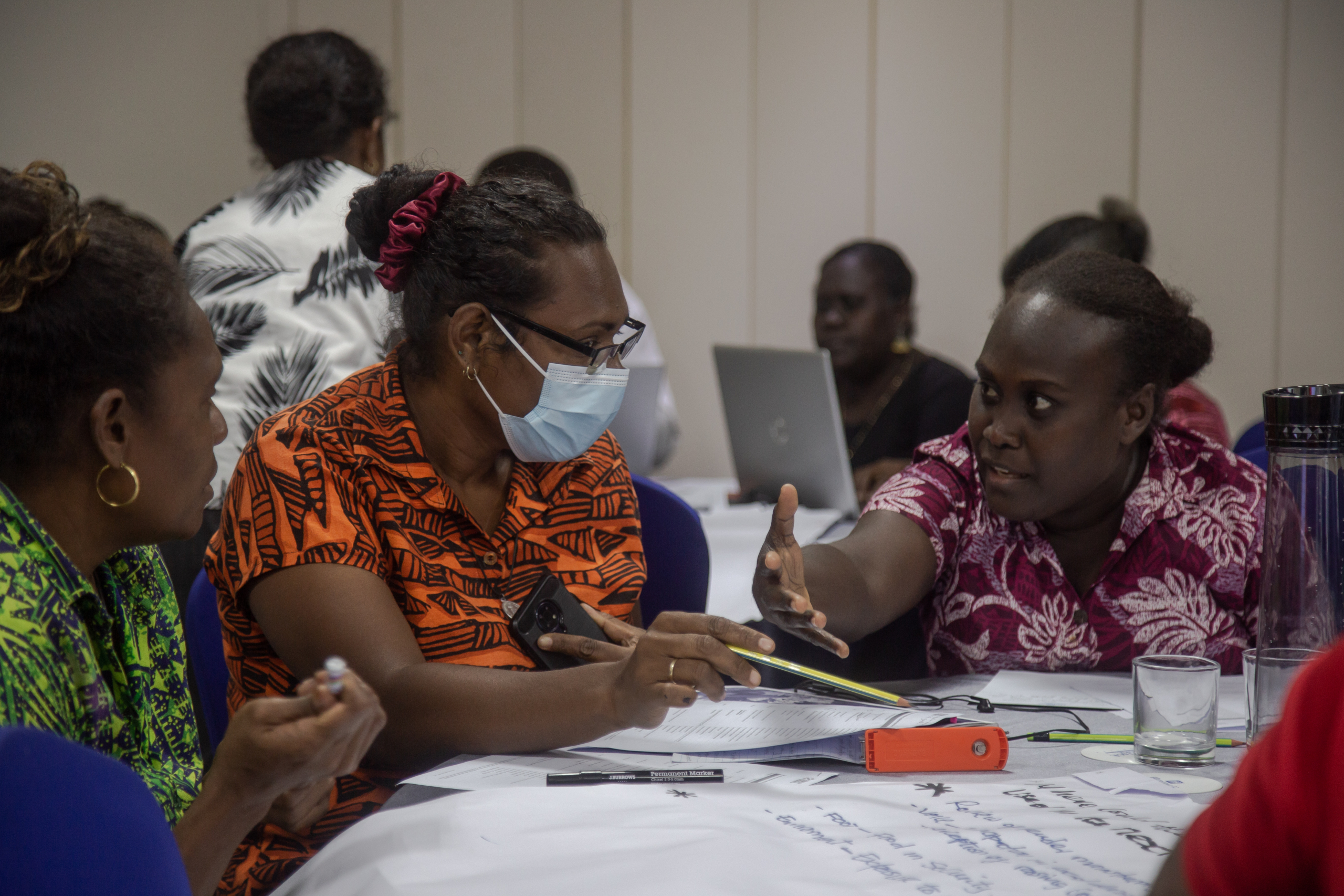 Three women are sitting around a desk in deep conversation. They are participating in gender data training in Solomon Islands. One is wearing a face mask.