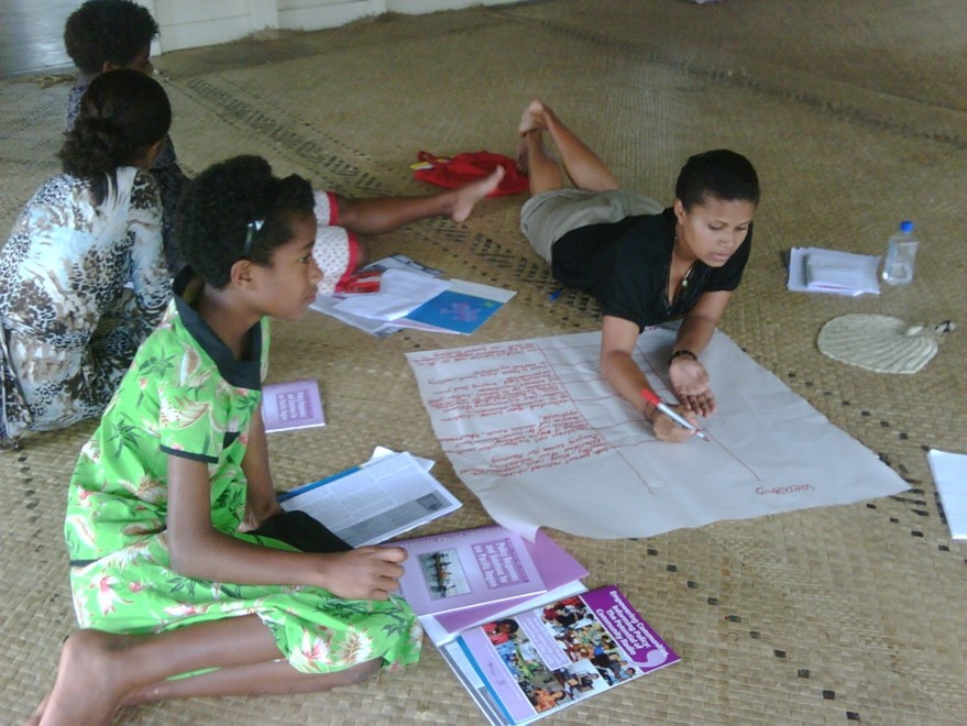A group of women in Fiji are sitting around a large piece of paper and other printed documents. They are writing on the paper and talking to each other.