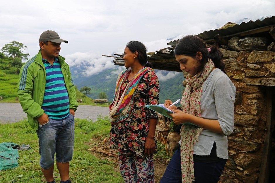An enumerator is surveying two people in Nepal. The enumerator is a women and she is writing on a a clip board. A man and a woman stand next to her answering questions.