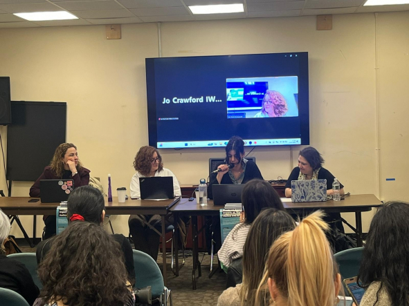 Four women sit at a table, one with a microphone, addressing a room. A screen behind them says Jo Crawford, IWDA