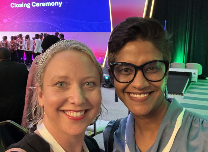 Two women stand smiling in front of a stage with the text Closing Ceremony in white text on a purple and blue screen.