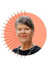 Smiling older woman with short gray hair wearing a silver chain necklace, portrait against a peach circular background with orange sunburst rays.